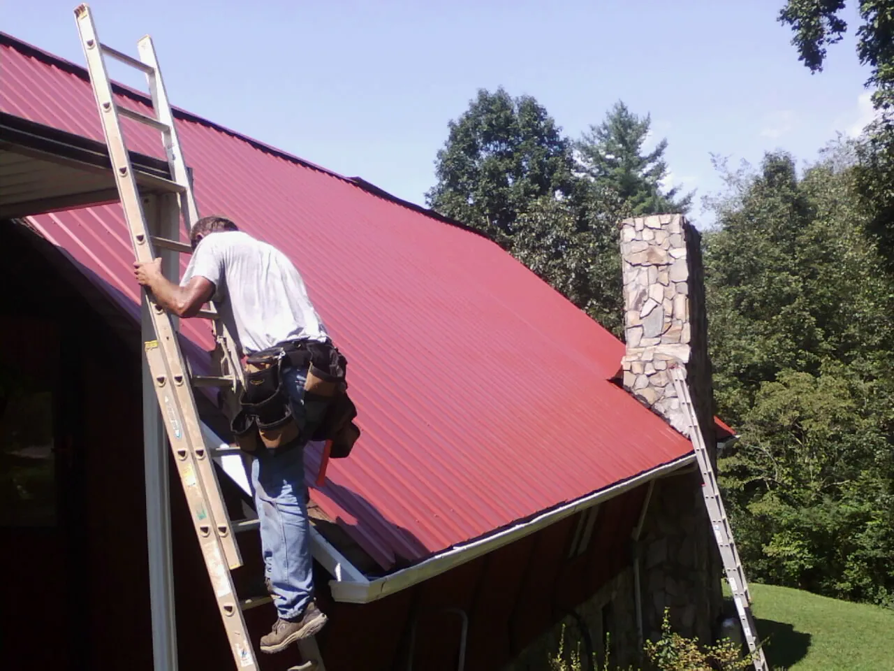 Roofer on a ladder beside a red metal roof and stone chimney — Soffit & Fascia Repair by Roofing Brothers in Port Byron